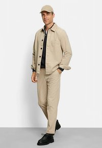 Beige Cordjacke aus Cord mit einer Fronttasche, kombiniert mit passenden Hosen. Schwarzes Hemd darunter und eine beigefarbene Kappe. Schwarze Schuhe.