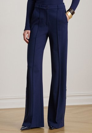 Kvinde iført marineblå bukser med vide ben, frontplisseringer og knapper i siden, kombineret med navyblå top og armbånd i guldfarve.