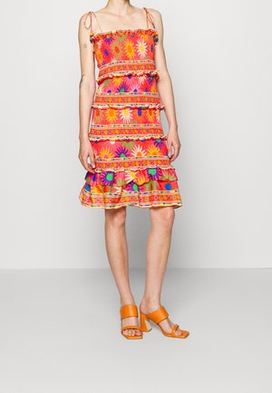 Femme portant une robe rose à fleurs sans manches avec des volants et des sandales à talons orange, debout devant un fond blanc uni.