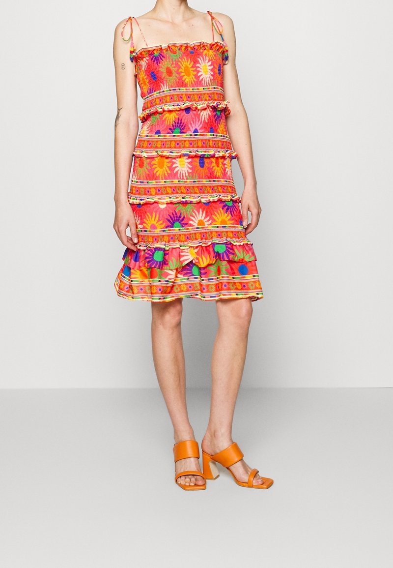 Femme portant une robe rose à fleurs sans manches avec des volants et des sandales à talons orange, debout devant un fond blanc uni.