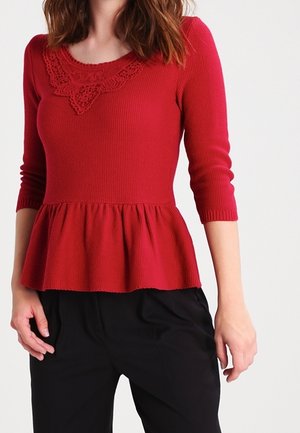 Červený pletený top s peplum střihem, zdobený krajkovým výstřihem a rukávy nad lokty. Textura je žebrovaná a silueta je přiléhavá v oblasti těla.