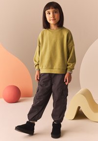 Sudadera verde texturizada con hombros caídos, combinada con pantalones cargo gris oscuro y botas negras hasta el tobillo. Fondo de color audaz con formas.