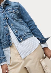 Džínová bunda ve světle modré barvě, s kapsami, stříbrnými knoflíky a roztrženými manžetami, vrstvená přes modro pruhovanou košili a béžové kalhoty.