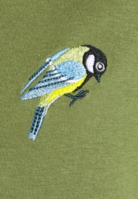 Broderad blå, gul, svart och vit fågel på grön tyg, detaljerad sömnad framhäver fjädrar och ansiktsdrag.