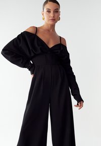 Moteris vilkinti juodą kombinezoną be petnešų su ilgomis rankovėmis ir auksiniais spiralės auskarais prieš paprastą baltą foną.