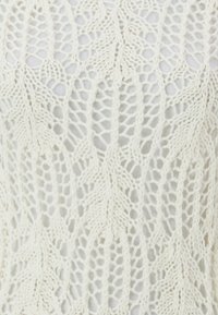 Koronkowa bluzka w kolorze ecru, zdobiona wzorem liści z ażurowym wykończeniem, prezentująca teksturowaną, przewiewną tkaninę.