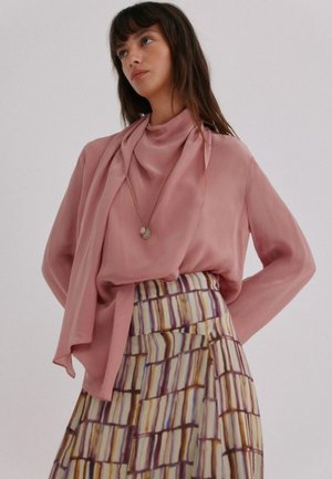 Vrouw met lang bruin haar draagt een rozenpinken blouse, een gouden hanger ketting en een geplooide rok met een meerkleurig abstract gestreept patroon.