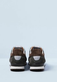 Ett par mörkgröna och bruna mockaskor med vita detaljer, sedda från baksidan på en ljusblå yta.