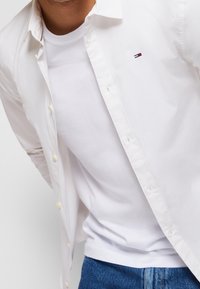White button-up shirt with yellow buttons over a white t-shirt. The shirt has a small red and blue logo on the chest and is paired with denim jeans.