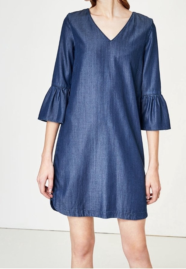 Džínové šaty s výstřihem do V, tříčtvrtečními rukávy a rozšířenými manžetami. Hladká textura, námořnická modrá barva a délka po kolena.