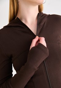 Sudadera con cremallera marrón, con capucha, diseño ajustado, mangas largas y agujeros para los pulgares. Textura suave y costuras visibles.