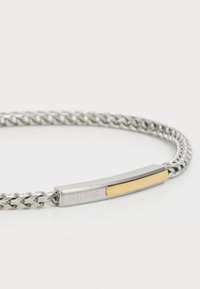 Bracelet en chaîne argentée avec plaque métallique rectangulaire dotée d'une bande dorée et du logo gravé "GUESS".