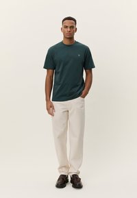 Zelena pamučna majica s okruglim izrezom i kratkim rukavima, uparena s kremasto obojenim hlačama i smeđim cipelama. Prisutan je detalj jednostavnog logotipa.