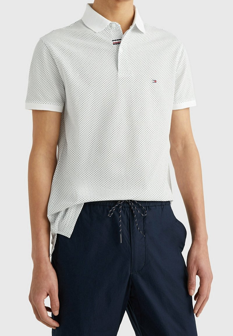 Polo shirt blanc à manches courtes avec de petits pois noirs, un col, et un petit logo rouge, blanc et bleu sur un pantalon de survêtement bleu marine à cordon.