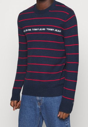 Man wearing navy blue sweater with red horizontal stripes and "Tommy Jeans" text across the chest, paired with blue jeans.