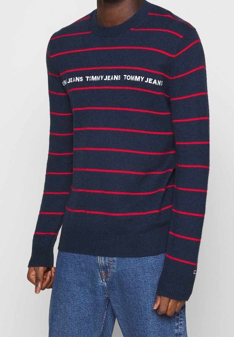 Homme portant un pull bleu marine avec des rayures horizontales rouges et le texte « Tommy Jeans » sur la poitrine, associé à un jean bleu.
