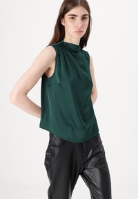 Satin groene mouwloze blouse met hoge halslijn en een relaxte pasvorm, gecombineerd met zwarte leren broek met zijzakken en een gladde textuur.