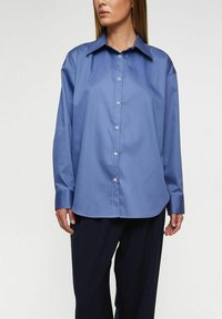 Ljusblå skjorta med knappar framtill, stor krage, långa �ärmar och slät textur. Har en avslappnad passform och subtil glans.