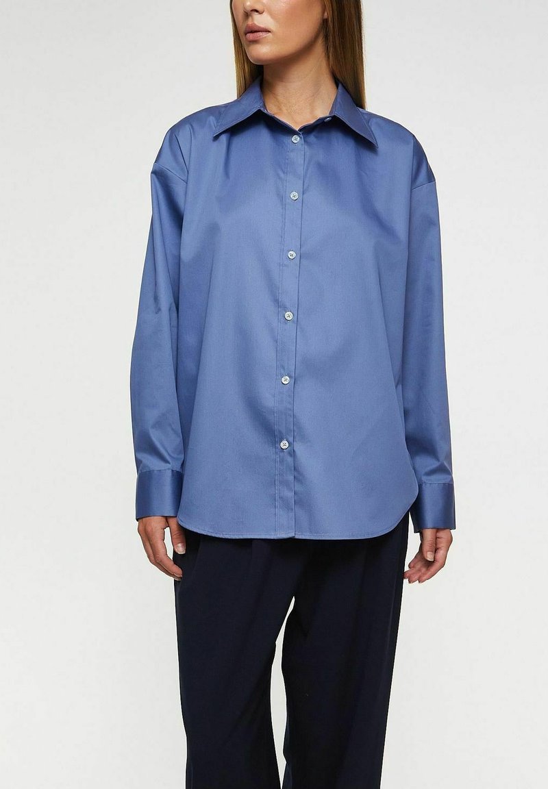 Ljusblå skjorta med knappar framtill, stor krage, långa ärmar och slät textur. Har en avslappnad passform och subtil glans.