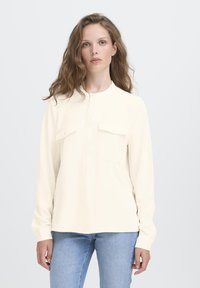 Kvinde med bølget hår iført en cremet knap-up skjorte med brystlommer og lyseblå jeans, stående foran en ensfarvet hvid baggrund.