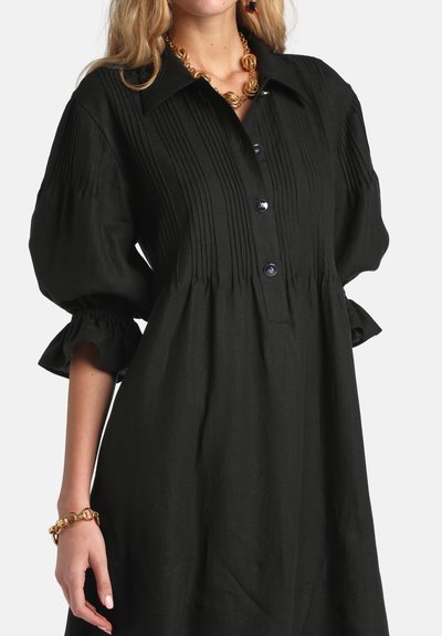 Woman wearing black buttoned dress with puffed three-quarter sleeves and pleated detailing, accessorized with gold chain necklace and bracelet.
