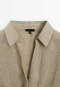 Col blanc en lin beige avec devant ouvert, montrant la texture du tissu et l'étiquette de la marque cousue à l'intérieur du col.