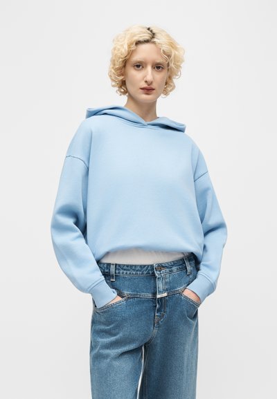 Άτομο με κοντά, κατσαρά ξανθά μαλλιά, ντυμένο με ανοιχτό μπλε φούτερ με κουκούλα και φαρδιά τζιν παντελόνια, με τα χέρια στις τσέπες, μπροστά σε απλό φόντο.