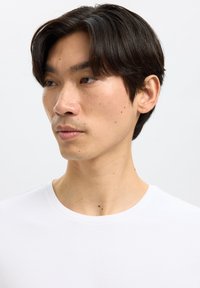 Jonge man met donker haar die een wit shirt met ronde hals draagt en naar links kijkt tegen een effen lichte achtergrond.