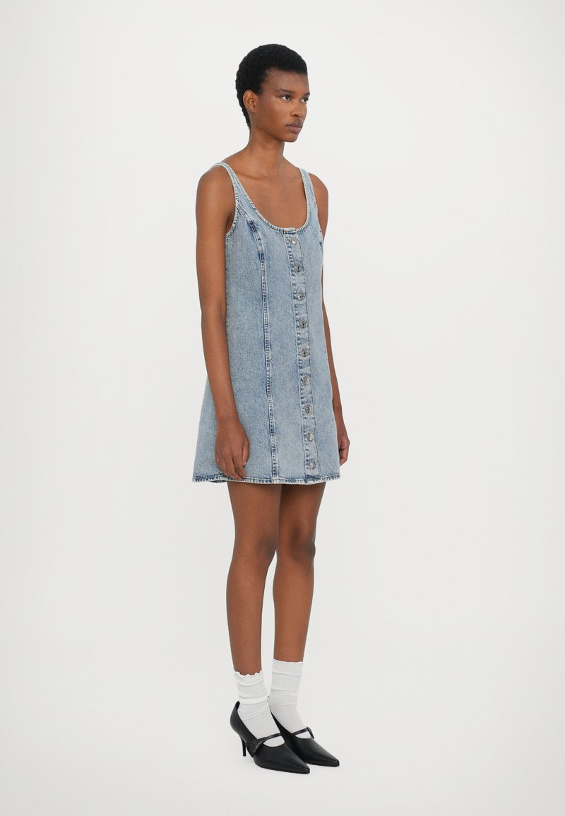 Jeanskleid mit verstellbaren Trägern, hat eine Knopfleiste vorne, eine ausgestellte A-Linie und eine hellblaue Wäsche. Getragen mit schwarzen Absätzen und weißen Socken.