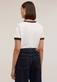 C�ămașă albă cu mâneci scurte, având un tiv ondulat burgund și accente albastre. Asortată cu pantaloni de blugi închis la culoare și o etichetă maro.