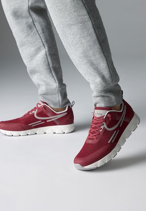 Personne portant un pantalon de survêtement gris et des baskets rouges avec des semelles blanches, debout sur une surface grise unie avec un fond gris.