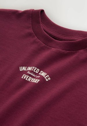 Bordo medvilninė marškinėlė su apvalia iškirpte, ant kurios baltomis raidėmis užrašyta „NEAPRIBOTOS ŠYPSENOS KASDIEN“ išlenktu dizainu.