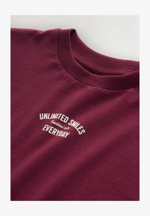 Vīna sarkanā kokvilnas T-krekls ar apaļu izgriezumu, uz kura ir uzdrukāts balts teksts "NEIEROBEŽOTAS SMAIDI KATRU DIENU" izliekta dizaina formā.