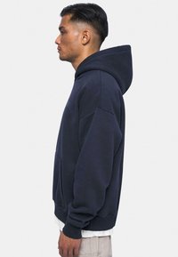Marineblå hættebluse med en frontlomme, oversized pasform og tekstureret stof. Har snøre i hætten og ribbede manchetter ved ærmerne.