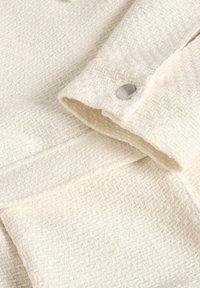 Kerman kangasta, jossa on teksturoitu kudonta, ja hihassa hopeinen nappi. Suunnittelussa on taitettu kaulus ja siistit linjat.