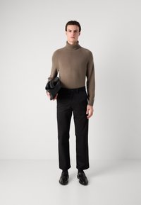 Strik turtleneck sweater i lys brun, parret med sorte bukser. Modellen holder en sort taske og bærer sorte sko, sat mod en neutral baggrund.