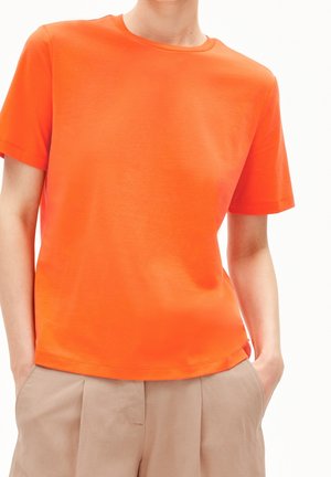 Jasnopomarańczowy bawełniany t-shirt z okrągłym dekoltem, krótkimi rękawami i luźnym krojem, w parze z beżowymi spodniami z bocznymi kieszeniami.