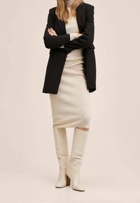 Robe en tricot crème avec des bottes en cuir blanches jusqu'aux genoux et un blazer noir, présentant une silhouette ajustée et une texture côtelée.