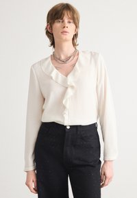 Hvid bluse med en flæsekantet V-udskæring, lange ærmer, båret med højtsiddende sorte jeans med små nitter for tekstur.