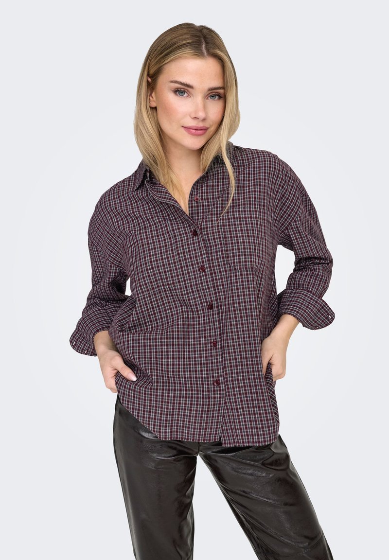 Chemise à carreaux rouge et bleu marine avec une coupe décontractée, manches longues, et un détail poche sur le côté gauche. Associée à un pantalon noir.