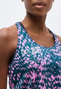 Κοντινό πλάνο ενός ατόμου που φοράει αθλητικό χωρίς μανίκια με πράσινο και ροζ αφηρημένο σχέδιο και λευκό λογότυπο στο στήθος.