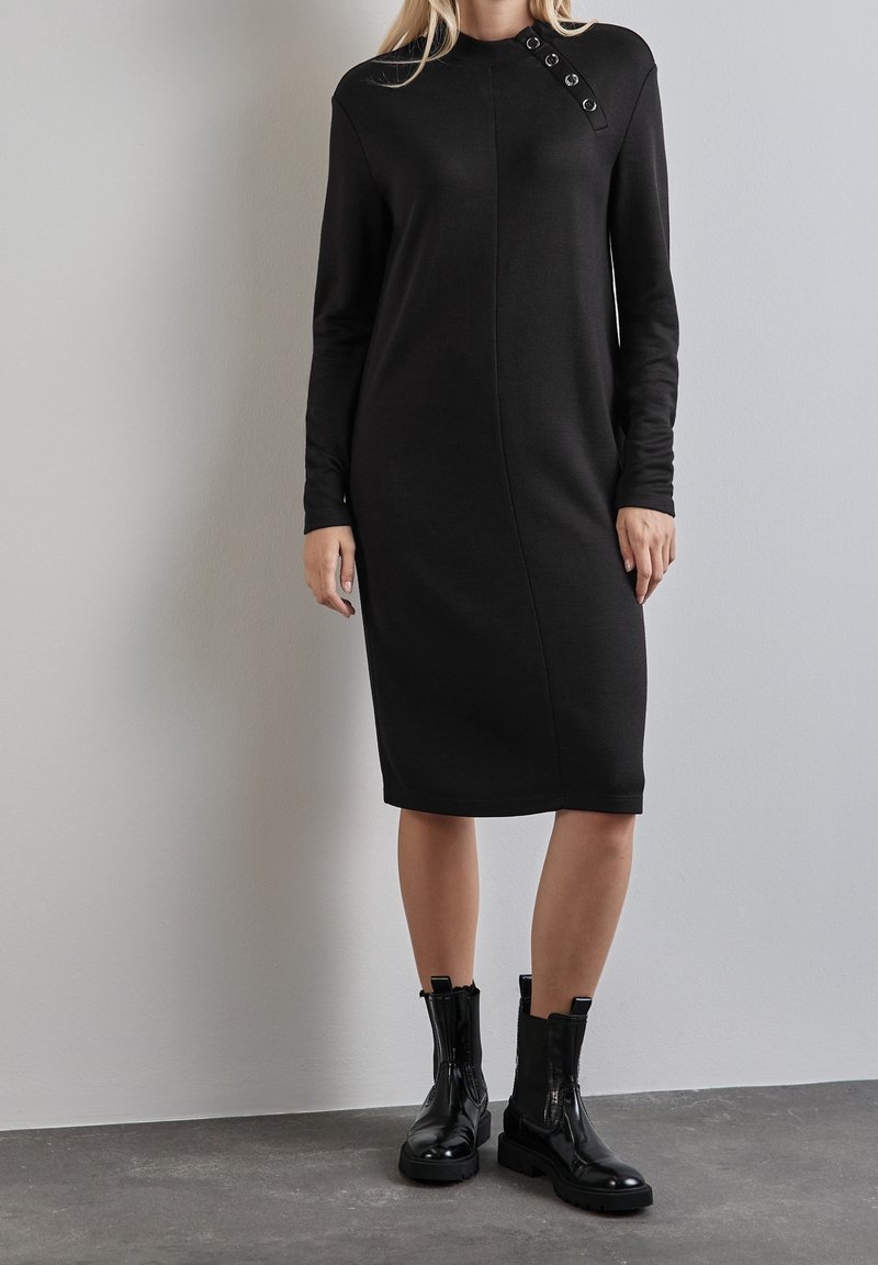 Femme portant une robe midi noire à manches longues avec des détails boutonnés sur l'épaule et des bottines noires, debout contre un mur gris.