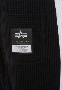 Crna teksturirana tkanina s prednjim džepom koji ima Alpha Industries Studio Edition oznaku s logotipom i podacima o proizvodnji.