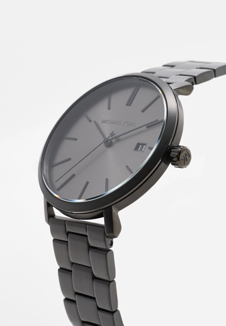 Montre-bracelet en métal noir Michael Kors avec un cadran simple, des marqueurs d'heure fins, un affichage de la date et un bracelet maillon sur fond blanc.