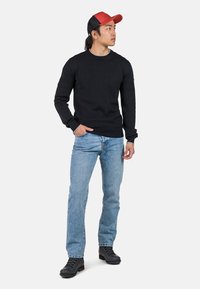 Schwarzer gestrickter Pullover mit langen Ärmeln, kombiniert mit hellblauen Jeans und schwarzen Schnürstiefeln. Eine rote Mütze rundet das Outfit ab.