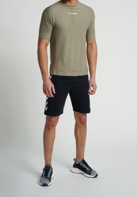 Man som bär en olivgrön kortärmad träningsskjorta, svarta shorts med vita chevronränder och gråsvart löparskor stående på vit golv.