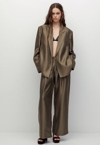 Completo verde oliva in satin con blazer oversize e pantaloni a gamba larga. Il blazer presenta rever a lancia, mentre il tessuto ha una texture liscia e lucida.