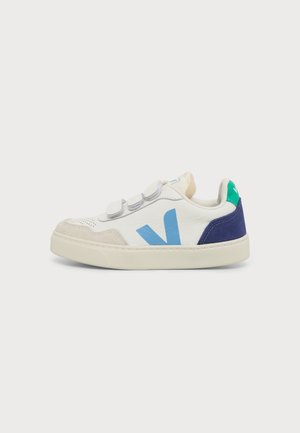 Zapatilla blanca de caña baja con detalles en azul y verde, suela beige y tres tiras de velcro grises.