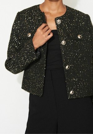 Personne portant une veste courte noire pailletée avec des boutons argentés sur un haut noir sans manches et un pantalon noir taille haute.