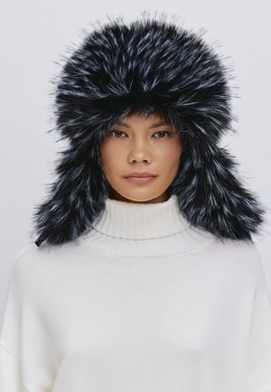 Căciulă din blană sintetică neagră, cu o coroană completă și clape pentru urechi, având o textur�ă moale. Purtați cu un pulover alb cu guler înalt din tricot.